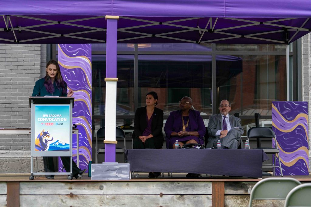 A speaker stand at a podium on an outdoor stagewith three other people sitting on the stage listening to the speaker.
