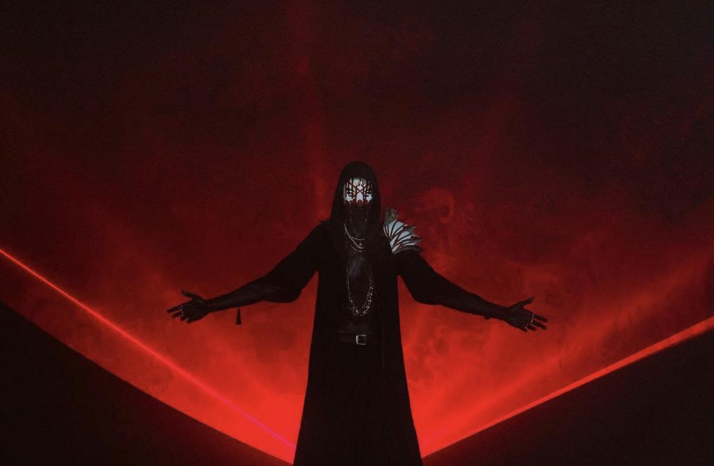 Black robed and masked figure standing with arms outspread in front of a red beam of light.