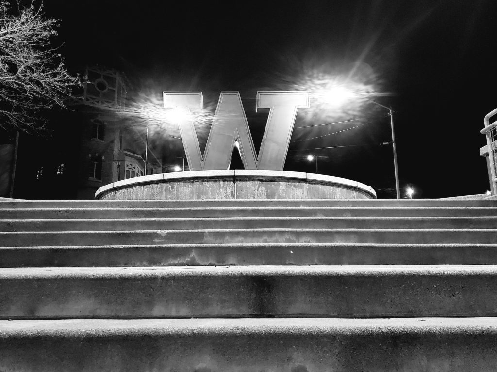 A black-and-white photo of a large W atop of concrete stairs, at night, with bright points of light backlighting the W.