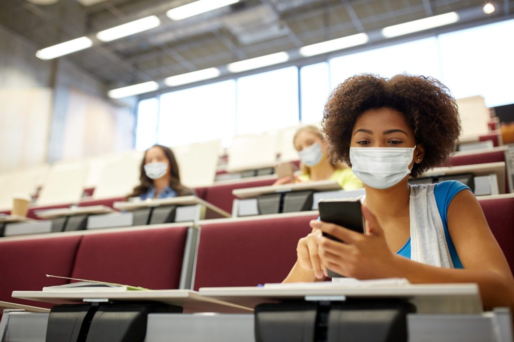 A young woman sits behind a desk, in a classroom, looking at her phone, while wearing a facemask and sitting at a distance from other blurry, masked, students.